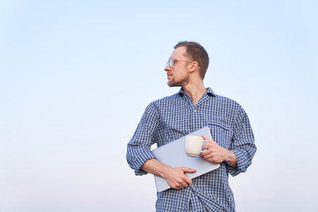 Cheerful bearded caucasian male staying outdoor in sleep wear with laptop and cup of coffee or tea. Clear blue sky at background. High quality imageの写真素材