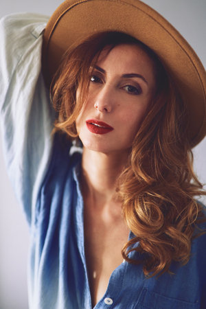 Posing perfectly looking young caucasian female in hat and jeans shirt with beautiful hair smiling looking at camera on grey background. High quality vertical photoの写真素材