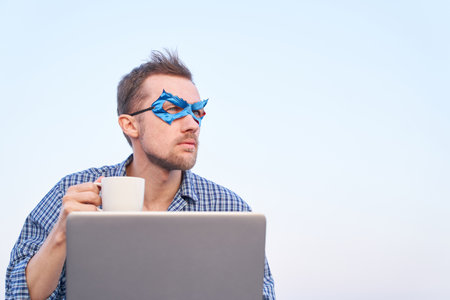 IT support superhero guy using laptop holding cup of hot tea or coffee. Cheerful man in nightwear shirt and blue face mask using laptop outdoors. High quality imageの写真素材
