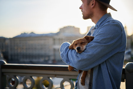 Dachshund puppy sleeping in the hands of the male owner. Caucasian man in denim shirt and baseball hat standing outside in the city with small puppy in hands relaxing or tired. Sunset timeの写真素材