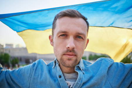 Closeup portrait of caucasian male in denim shirt with flag of Ukraine waving on wind with urban view on background. Patriot man with flag of Ukraine. Freedom concept. High quality photoの写真素材