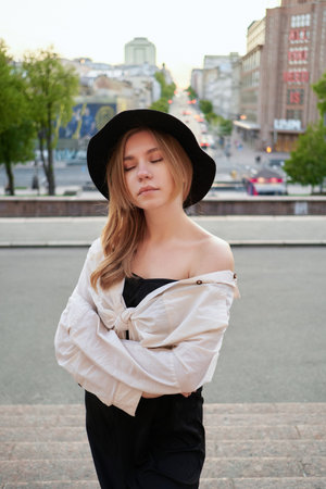 Adorable natural looking caucasian blonde teenage girl in black dress and hat posing outdoor with urban view background at sunset. Young hipster portrait. High quality vertical photoの写真素材