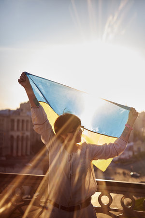 Caucasian female silhouette in white blouse with flag of Ukraine waving on wind with urban view on background and lens flares effect. Portrait of a patriotic woman with flag of Ukraine. Freedom timeの写真素材