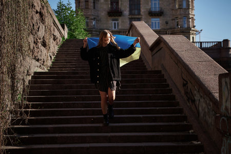 Adorable caucasian girl in jeans jacket running down on staircase holding flag of Ukraine. The concept of freedom or patriotism. Girl with the blue-yellow Ukrainian flag. High quality imageの写真素材