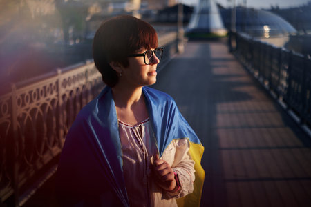 Confidence caucasian girl in white blouse and eyeglasses covered with a flag of Ukraine looking at side. Portrait of patriotic woman with flag of Ukraine at sunset. Freedom concept. High quality photoの写真素材