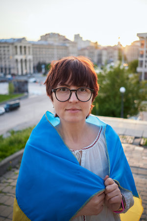 Caucasian girl in white blouse and eyeglasses covered with a flag of Ukraine against of a city view. Portrait of a patriotic woman with flag of Ukraine at sunset. Freedom concept. High quality photoの写真素材