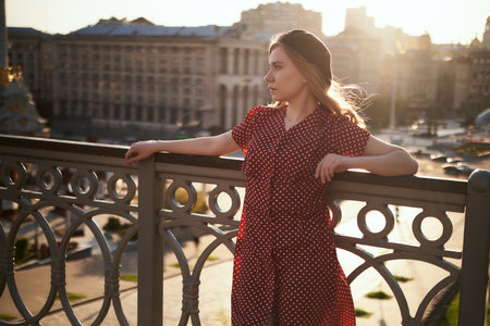 Fashion portrait of adorable stylish blonde young caucasian girl in beret and polka dot dress posing outside looking at camera with urban view and lens flares effect at sunset. High quality imageの写真素材