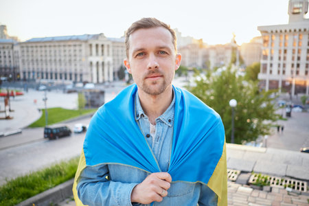 Caucasian male in denim shirt covered with a flag of Ukraine against of a city view. Portrait of a patriot man with flag of Ukraine. Sunset time. Freedom concept. High quality photoの写真素材