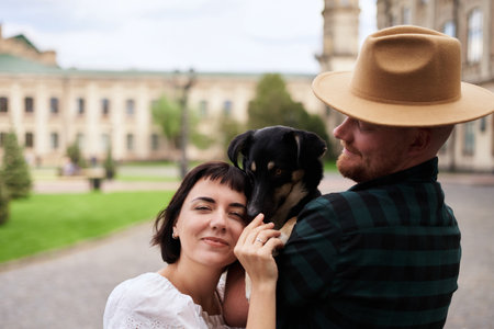 Happy couple with a shelter dog hugging in background of the old city in summer. Women with black hair and man in a hat, cute young modern couple with a rescued dog. High quality photoの写真素材