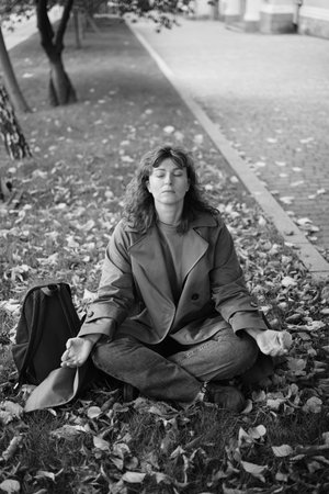 Caucasian middle aged woman meditating in lotus pose at autumn city street with leaves. Woman relaxing on yellow leaves. Meditation, Mental health, self care, mindfulness. High quality photoの写真素材