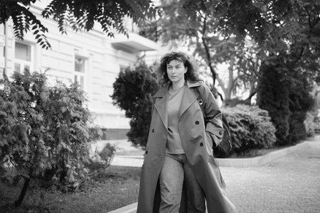 Young adult trendy stylish beautiful caucasian casual woman walking the urban city street with backpack. Female person portrait wear jeans brown trench coat in hurry to meeting. High quality photoの写真素材