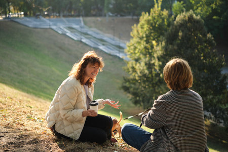 Two friends talking and laughing sitting on the grass in a park. Drinking coffee to go, with small dog in the sweater. Sunny evening light, weekend concept. High quality photoの写真素材