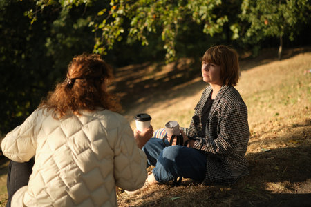 Two friends talking and laughing sitting on the grass in a park. Drinking coffee to go, with small...