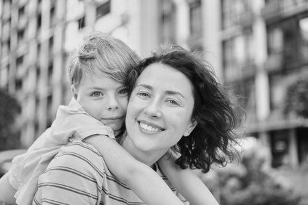 Outdoor family day concept. Portrait of adorable caucasian brunette mother sitting and kid son hugging her from the backの写真素材
