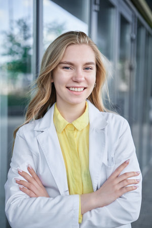 Positive female medical student in white uniform standing near clinic entrance and looking at camera smiling. Beautiful young blond doctor, veterinarian High quality photoの写真素材