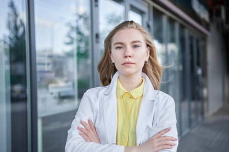 Positive female medical student in white uniform standing near clinic entrance and looking at camera smiling. Beautiful young blond doctor, veterinarian High quality photoの写真素材