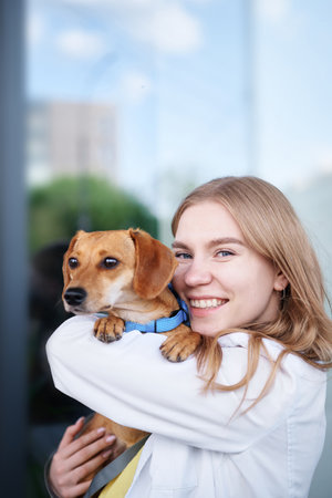 Young veterinarian smiling and holding dog near the vet clinic entrance. Healthy dog, diagnostic, treatment of domestic animals. High quality photoの写真素材