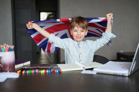 happy boy smiling and holding British flag sitting zt desk with laptop and notes. Concept of English learning at home High quality photoの写真素材
