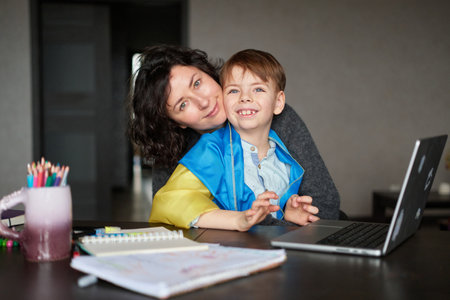 Ukrainian mother and son with flag sitting with laptop at home studying .Happy smiling family hugs and looking at the camera. Ukrainian refugees concept. High quality photoの写真素材