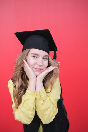 Young beautiful blond female in graduation cap with emotional facial expression on red background. Happy to finish school, college or university High quality photoの写真素材