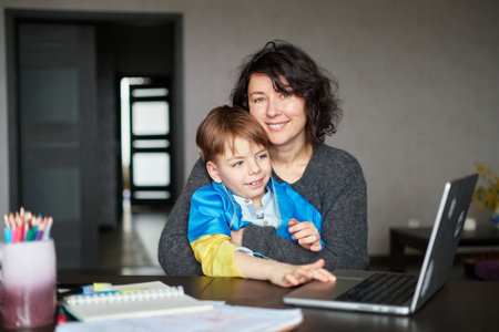 Ukrainian mother and son with flag sitting with laptop at home studying .Happy smiling family hugs and looking at the camera. Ukrainian refugees concept. High quality photoの写真素材