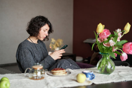Positive middle age women sitting the the table at home with tea and pancakes. Using smartphone . Beautiful lade looking at the camera, smiling, wearing casual clothing. High quality photoの写真素材