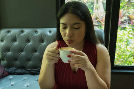 Woman in red dress holding hot coffee cup and blowing coffee to make hot coffee cold in coffee shop day time with relaxing mood and happy faceの写真素材