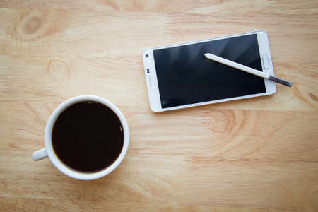 Mobile phone with stylus pen and coffee on wooden table.