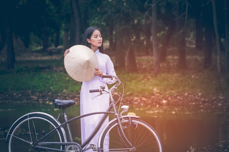 Portrait of beautiful young Asian teen with long hair wearing a costume of Viet Nam (Ao dai) she is currently towed a bicycle and holding a hat by verdant gardens background with copy space.の写真素材