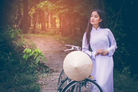 Portrait of beautiful young Asian teen with long hair wearing a costume of Viet Nam (Ao dai) she is currently towed a bicycle and holding a hat by verdant gardens background with copy space.の写真素材