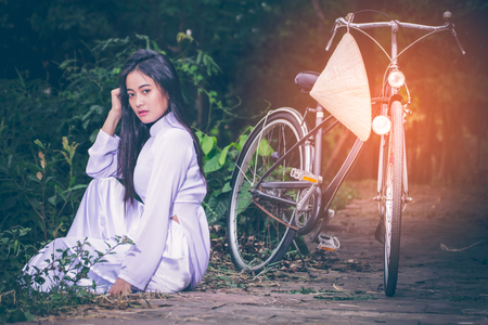 Portrait of young Asian girl with long hair wearing a dress of Vietnam (Ao Dai) sitting beside her bike, and hats woven palm leaves hanging on bicycle handlebars. Set amidst the lush gardens with copy space.の写真素材