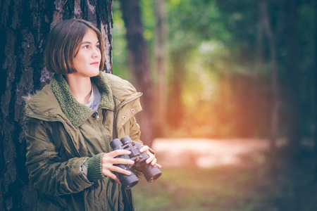 Beautiful girl teen American hikers looking up on the tree. She wore wearing a green sweater with binoculars in the hand. In the warm sunlight in the forest with copy space.の写真素材
