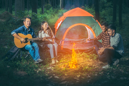 A group of tourists trekking Asians sitting in front of the tent all while relaxing camping by playing the guitar and made a fire from the firewood within the pine forest and the night atmosphere.の写真素材