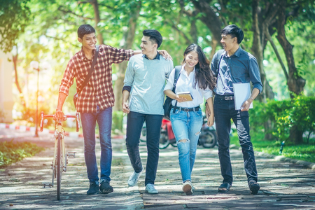 A group of Asian student was walking on the road within the University. They talk to smile happily and motivate the bicycle to walk together. In the shade of trees streets and the sun in the afternoonの写真素材