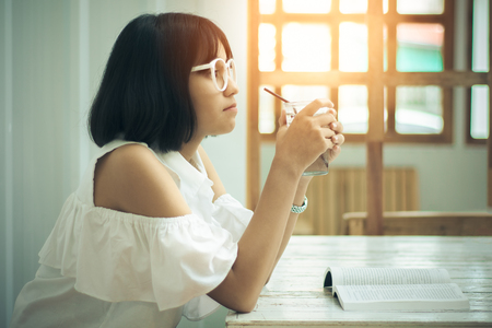 Asian teen girls wearing white shirts and glasses. She is holding a glass of pure water in her hand, showing emotions, thought and vision by reading the books, which are placed on the white table.の写真素材