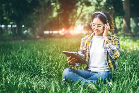 Asian hipster curly girl sitting on the lawn She wears headphones and smiles happily while using the tablet to relax and study online. in the atmosphere of the soft sunlight in the morning.の写真素材