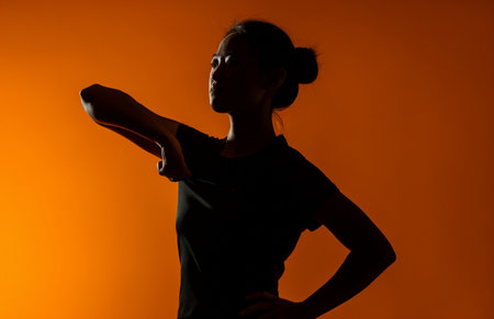 Silhouette of young Asian woman stretching her arms on orange backgroundの素材