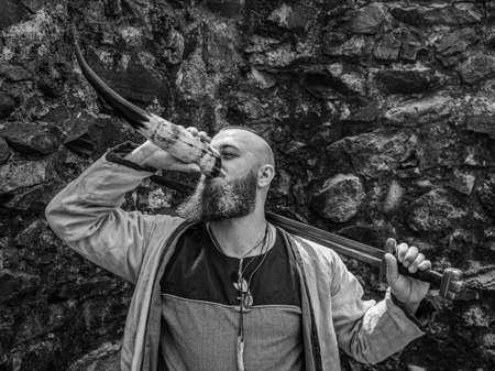 Viking warrior with thick beard and big sword drinks from the horn in front of a stone wall, black and white historical reenactment imageの写真素材
