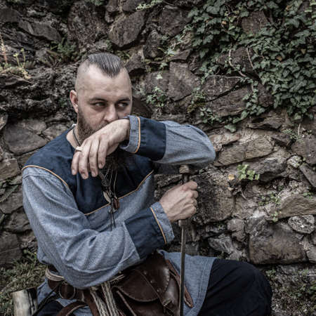 Viking warrior with thick beard sits thoughtfully in front of a stone wall, image of historical...