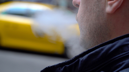 Young Millennial urban hipster blowing vapor electronic cigarette smoke outside on New York City street in day time. Yellow taxi cab passes in background. Over the shoulder POV viewの写真素材