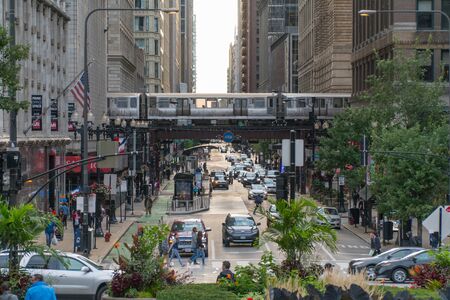 Chicago, IL, USA - Circa 2019: View down busy street in downtown as Loop train passes overhead tracks over avenue morning rush hour traffic and crowds of people walking down sidewalks to workの写真素材