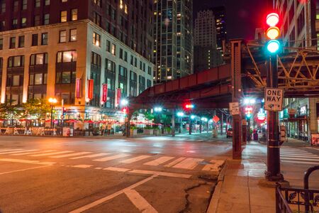 Chicago, IL - Circa 2019: Night time long exposure of busy downtown intersection under loop train tracks with traffic control light lit green yellow red for vehicle safety driving on busy roadway through city.の写真素材