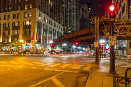 Chicago, IL - Circa 2019: Night time long exposure of busy downtown Chicago intersection roadway under loop train track. Red traffic light on corner directs vehicle and pedestrians across crosswalk for safetyの写真素材