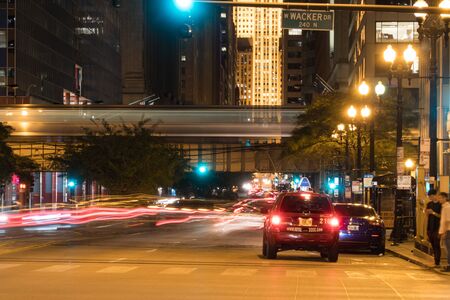 Chicago, IL - Circa 2019: Night time long exposure downtown as busy car traffic drives through intersection past pedestrian crossing people waiting at light to safely cross roadway during rush hour.の写真素材