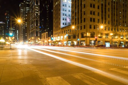 Night time long exposure of busy urban road way driving through urban city area past skyscrapers downtown with light trails of cars crossing intersection crosswalkの写真素材
