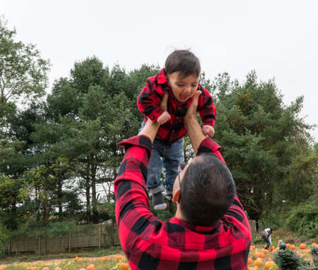 A proud father lifts his infant son in the air outside during fall autumn season to bring joy and happiness to child laughing and smiling faceの写真素材