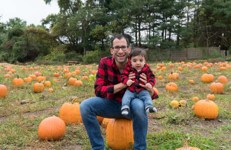Handsome father and adorable infant son pose for autumn holiday portraits sitting on large pumpkin in a field wearing matching plaid button down flannel shirtsの写真素材