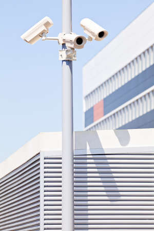 Three surveillance cameras on the pole, white building and sky in background