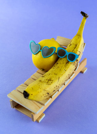 Lemon and banana with sunglasses resting on the wooden beach bed. Summer vacation concept on purple backgroundの写真素材