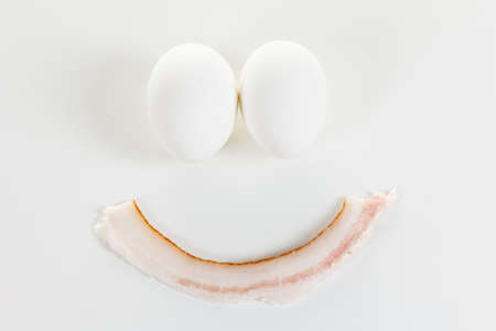 Two chicken eggs and a slice of bacon in the form of a smile on a white background. Humor conceptの写真素材